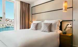 Hotellihuone, jossa on parisänky, koristetyynyt ja yöpöytä. Näkymä vedelle ja rakennuksiin ikkunan kautta. (Tekoälyn luoma teksti)