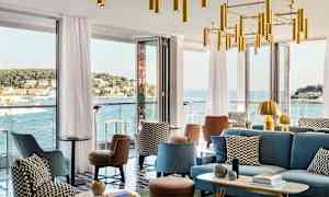 Moderni lounge, jossa istuimia ja koristeellisia kattokruunuja, ympärillä suuret ikkunat, jotka tarjoavat näkymän rannikkokaupunkiin ja merelle. (Tekoälyn luoma teksti)