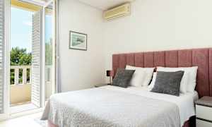 Vaalea makuuhuone, jossa on vaaleanpunainen sängynpääty, valkoinen päiväpeite ja ovi parvekkeelle. (Tekoälyn luoma teksti)