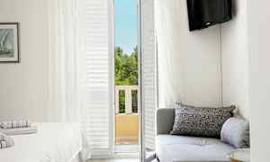 Hotellihuone, jossa sohva, sänky ja parvekeovi, josta avautuu näkymä puihin. (Tekoälyn luoma teksti)