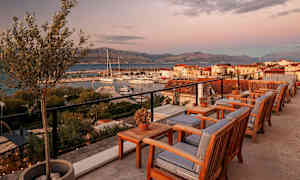 Ulkoterassi, jossa puisia tuoleja ja pöytiä, näkymä venesatamaan ja vuoristoon auringonlaskussa. (Tekoälyn luoma teksti)