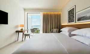 Hotellihuone, jossa on sijattu sänky, kirjoituspöytä, tuoli ja näkymä merelle. (Tekoälyn luoma teksti)