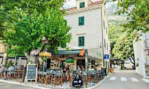 Ulkoilmakahvila, joka sijaitsee valkoisessa rakennuksessa vihreiden ikkunaluukkujen ympäröimänä, ympärillä vehreyttä ja puita. (Tekoälyn luoma teksti)