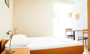 Yksinkertainen hotellihuone, jossa on puinen sänky, yöpöytä ja pöytä kahdella tuolilla. (Tekoälyn luoma teksti)