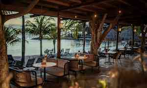 Ulkosisustusalue puukatoksen alla, jossa sohvia ja pöytiä meren äärellä, ympärillä palmupuita. (Tekoälyn luoma teksti)