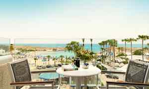 Näkymä parvekkeelta, jossa pöytä ja tuolit, rannalle, jolla on aurinkovarjoja ja palmuja meren äärellä. (Tekoälyn luoma teksti)