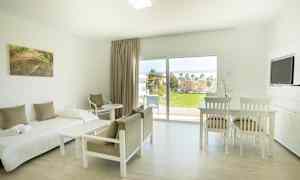 Vaaleasti sisustettu huone, jossa sänky, tuolit, pöytä ja näkymä viheralueelle suurten ikkunoiden kautta. (Tekoälyn luoma teksti)