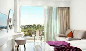 Vaalea hotellihuone, jossa sohva, sänky violetilla peitteellä ja parveke, jolta näkyy vehreää maisemaa. (Tekoälyn luoma teksti)