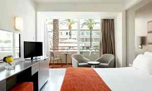 Hotellihuone, jossa sänky, istuinpaikat ja parveke näkymällä sisäpihalle palmuineen. (Tekoälyn luoma teksti)