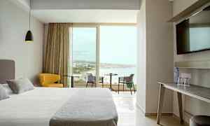 Hotellihuone, jossa on moderni sisustus, leveä sänky, keltainen tuoli ja pöytä, josta on näkymä rannalle suurten ikkunoiden kautta. (Tekoälyn luoma teksti)