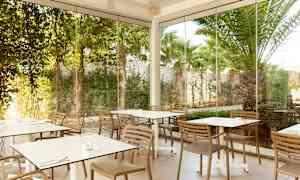 Kuva ulkoterassista, jossa on pöytiä ja tuoleja, joiden ympärillä on lasiseiniä ja vihreää kasvillisuutta. (Tekoälyn luoma teksti)