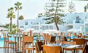 Ravintola, jossa on puisia tuoleja ja katettuja pöytiä, näkymä valkoiseen arkkitehtuuriin, altaalle ja palmuihin. (Tekoälyn luoma teksti)