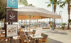 Ulkoravintolan pöytiä ja tuoleja suuren aurinkovarjon alla satamassa. (Tekoälyn luoma teksti)