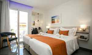 Valoisa hotellihuone, jossa on kaksi yksittäissänkyä pedattuna valkoisilla lakanoilla ja oransseilla koristetyynyillä. Huoneessa on parveke lasiovilla, tuoli ja peilipöytä. (Tekoälyn luoma teksti)