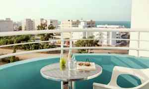 Parveke, josta on näkymä kaupungille ja merelle. Pöytä, jolla on lasi, vihreä omena, kirja ja aurinkolasit. (Tekoälyn luoma teksti)