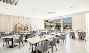 Valoisa ja moderni ravintola-alue, jossa on pyöreitä ja neliskulmaisia pöytiä pehmustettujen tuolien ympäröiminä. Suuret ikkunat tarjoavat näkymän ulkoalueelle. (Tekoälyn luoma teksti)