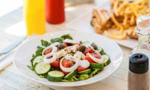 Lautasellinen salaattia, jossa on tomaattia, kurkkua, sipulia, fetajuustoa ja oliiveja. Taustalla näkyy ranskalaisia ja voileipiä. (Tekoälyn luoma teksti)