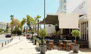 Kuva ulkoilmaravintolasta ja jalkakäytävästä kadun varrella, taustalla puita ja rakennuksia. (Tekoälyn luoma teksti)