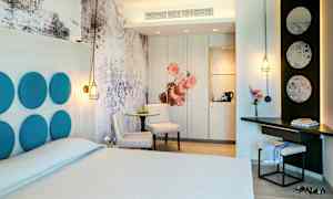 Hotellihuone modernilla sisustuksella, mukaan lukien parivuode, pöytä tuoleineen sekä yksilöllisiä yksityiskohtia kuten koristepaneeleja ja valaistusta. (Tekoälyn luoma teksti)