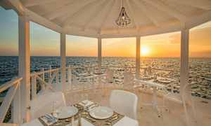 Ulkoravintola, jossa on valkoiset kalusteet, sijaitsee meren rannalla ja taustalla näkyy auringonlasku. (Tekoälyn luoma teksti)