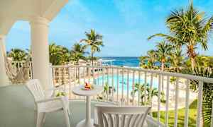 Kuvassa näkyy rentouttava parveke, jossa on pöytä ja tuoleja sekä riippumatto. Näkymä avautuu trooppiseen uima-altaaseen, jota ympäröivät palmut, ja taustalla on meri. (Tekoälyn luoma teksti)