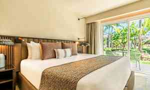 Hotellihuone, jossa on suuri sänky useilla tyynyillä, yöpöytä ja lasiovi, joka johtaa ulkoterassille ja vehreään ympäristöön. (Tekoälyn luoma teksti)