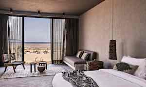 Hotellihuone, jossa sänky, sohva ja näkymä rannalle suuren lasioven kautta. (Tekoälyn luoma teksti)