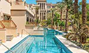 Pitkä kapea uima-allas palmujen ja arabialaista arkkitehtuuria edustavien rakennusten ympäröimänä. (Tekoälyn luoma teksti)