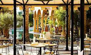 Ulkotarjoilualue, jossa on pöytiä ja tuoleja katoksen alla, ympäröitynä kasvillisuudella ja palmupuiden siimeksessä. (Tekoälyn luoma teksti)