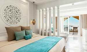 Hotellihuone, jossa on vaaleat seinät, koristeltu sänky sinisillä ja beigeillä vuodevaatteilla sekä parveke merinäköalalla. (Tekoälyn luoma teksti)