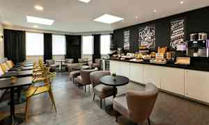 Kahvila-alue, jossa on pöytiä, tuoleja ja seinän vieressä penkki. Taustalla näkyy tarjoilupöytä, jossa on aamiaistarvikkeita ja kahvinkeitin. (Tekoälyn luoma teksti)