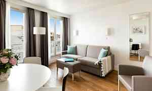 Moderni, valoisa olohuone, jossa harmaa sohva, pöytä, nojatuoli ja suuret ikkunat, joista tulee luonnonvaloa. (Tekoälyn luoma teksti)