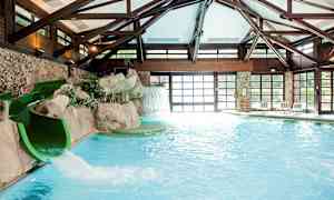 Sisäuima-allas, jossa on vesiliukumäki lähellä kallioita ja keinotekoista vesiputousta. Allasalueen ympärillä on suuria ikkunoita ja puurakenteita. (Tekoälyn luoma teksti)