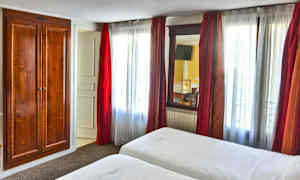 Hotellihuone, jossa on kaksi sänkyä, valkoiset lakanat ja puinen vaatekaappi ikkunan vieressä, jossa on punaiset verhot. (Tekoälyn luoma teksti)