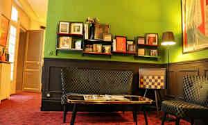 Huone, jossa on vihreät seinät ja eklektistä sisustusta, mukaan lukien hylly kirjoineen ja koristeineen, sohva ja shakkikuviollinen tuoli. (Tekoälyn luoma teksti)