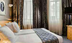 Hotellihuone, jossa kaksi erillistä vuodetta, vaaleat verhot ja koristeellinen sängynpäällinen. (Tekoälyn luoma teksti)