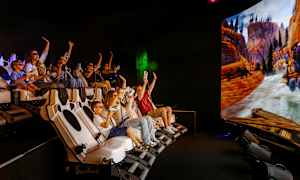 Ryhmä ihmisiä istuu interaktiivisessa simulaattorissa, jossa iso näyttö näyttää luontomaiseman vuorineen ja jokiineen. (Tekoälyn luoma teksti)