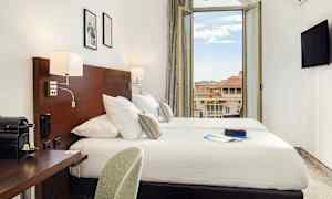 Hotellihuone, jossa on parivuode, kirjoituspöytä ja avonainen parvekkeen ovi, joka näyttää näkymän ympäröivistä rakennuksista. (Tekoälyn luoma teksti)