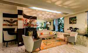 Olohuone, jossa on mukavat nojatuolit, pöytä ja sohva oranssilla matolla, sekä kasveja ja taidetta seinillä. Iso peili ja ikkunat. (Tekoälyn luoma teksti)