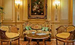 Tyylikkäästi sisustettu huone, jossa on koristeellinen sohva, kaksi sopivaa nojatuolia ja sohvapöytä edessä. Öljymaalaus sohvapöydän yläpuolella seinällä. (Tekoälyn luoma teksti)