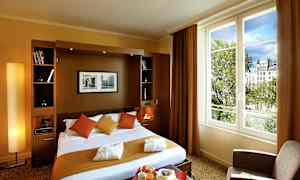 Hotellihuone, jossa on parisänky, tyynyjä ja peittoja lämpimissä sävyissä sekä suuri ikkunanäkymä puille ja rakennuksille. (Tekoälyn luoma teksti)