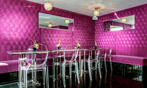 Moderni ja värikäs ravintola tai lounge, jossa on fuksian värinen pehmustettu seinäpaneeli, läpinäkyvät tuolit ja peilikoristeita. (Tekoälyn luoma teksti)