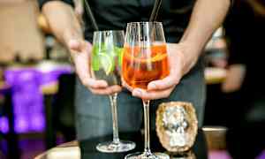 Henkilö pitelee kahta juomaa, joista toisessa lasissa on oranssi cocktail ja toisessa kirkas cocktail limeviipaleiden ja mintun kera. (Tekoälyn luoma teksti)