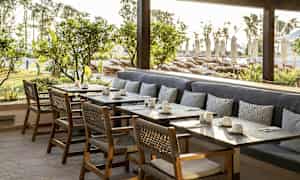 Ulkotila, jossa pöytiä, tuoleja ja istuinpenkki, taustalla vehreyttä ja aurinkotuoleja. (Tekoälyn luoma teksti)