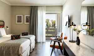 Hotellihuone, jossa kaksi erillistä vuodetta, istuinryhmä ja kirjoituspöytä. Parveke näkymällä puihin. (Tekoälyn luoma teksti)