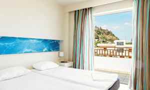Hotellihuone, jossa on kaksi erillistä vuodetta, vaaleat seinät ja sininen koristeellinen seinäpaneeli. Suuri ikkuna avautuu näkymään kukkulalle ja rakennuksille ulkona. (Tekoälyn luoma teksti)
