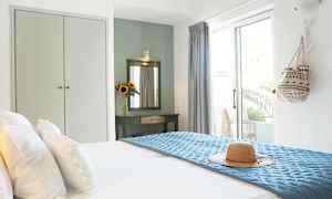 Valoisa hotellihuone, jossa on pedattu sänky, olkihattu sinisellä päiväpeitolla ja koristeellinen pöytä peilin kanssa sekä auringonkukkamaljakko. (Tekoälyn luoma teksti)