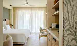 Hotellihuone, jossa on iso sänky, nojatuoli ja hyllyjä kahvinkeittimellä, valaistuna luonnonvalolla suurten verhojen läpi. (Tekoälyn luoma teksti)