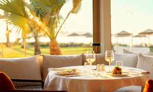 Pöytä katettu ravintolassa, ruokaa ja lasit aurinkoisen näköalan äärellä palmuineen. (Tekoälyn luoma teksti)