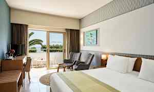 Hotellihuone, jossa on parivuode, kaksi tuolia, pöytä ja TV-huonekalu. Lasisen oven kautta näkyy parveke, josta on näkymä palmuihin. (Tekoälyn luoma teksti)